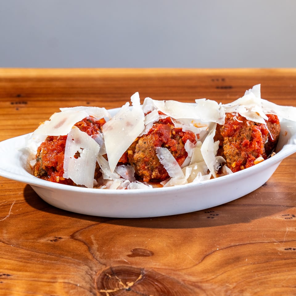 Meatballs covered in tomato sauce and shaved Parmesan served in a white dish on a wooden table.
