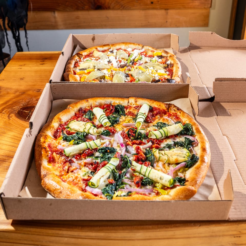 Two vegetable-topped pizzas in open cardboard boxes on a wooden table, featuring zucchini ribbons, leafy greens, peppers, onions, and cheese.