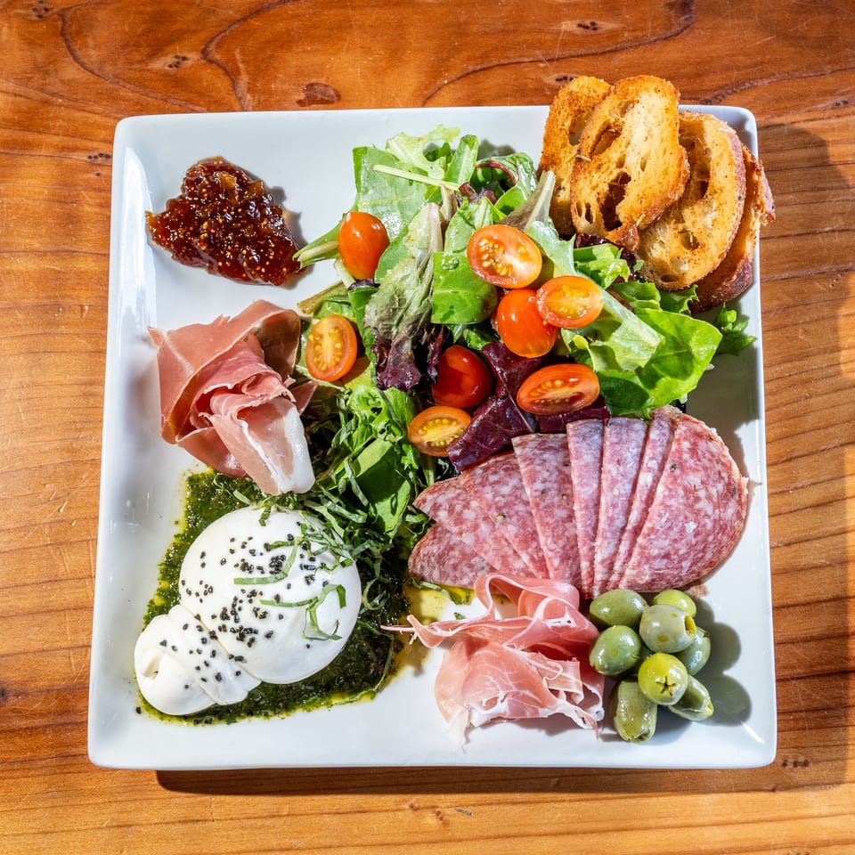 Antipasto platter with mixed greens, tomatoes, cured meats, burrata, olives, toasted bread, pesto, and fig jam on a wooden table.