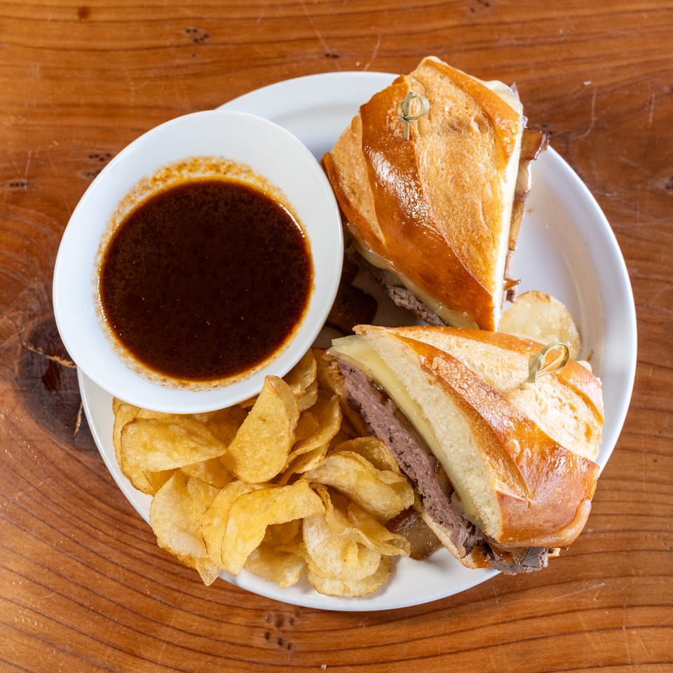 Plate of toasted roast beef sandwiches, potato chips, and a bowl of dark dipping sauce on a wooden table.